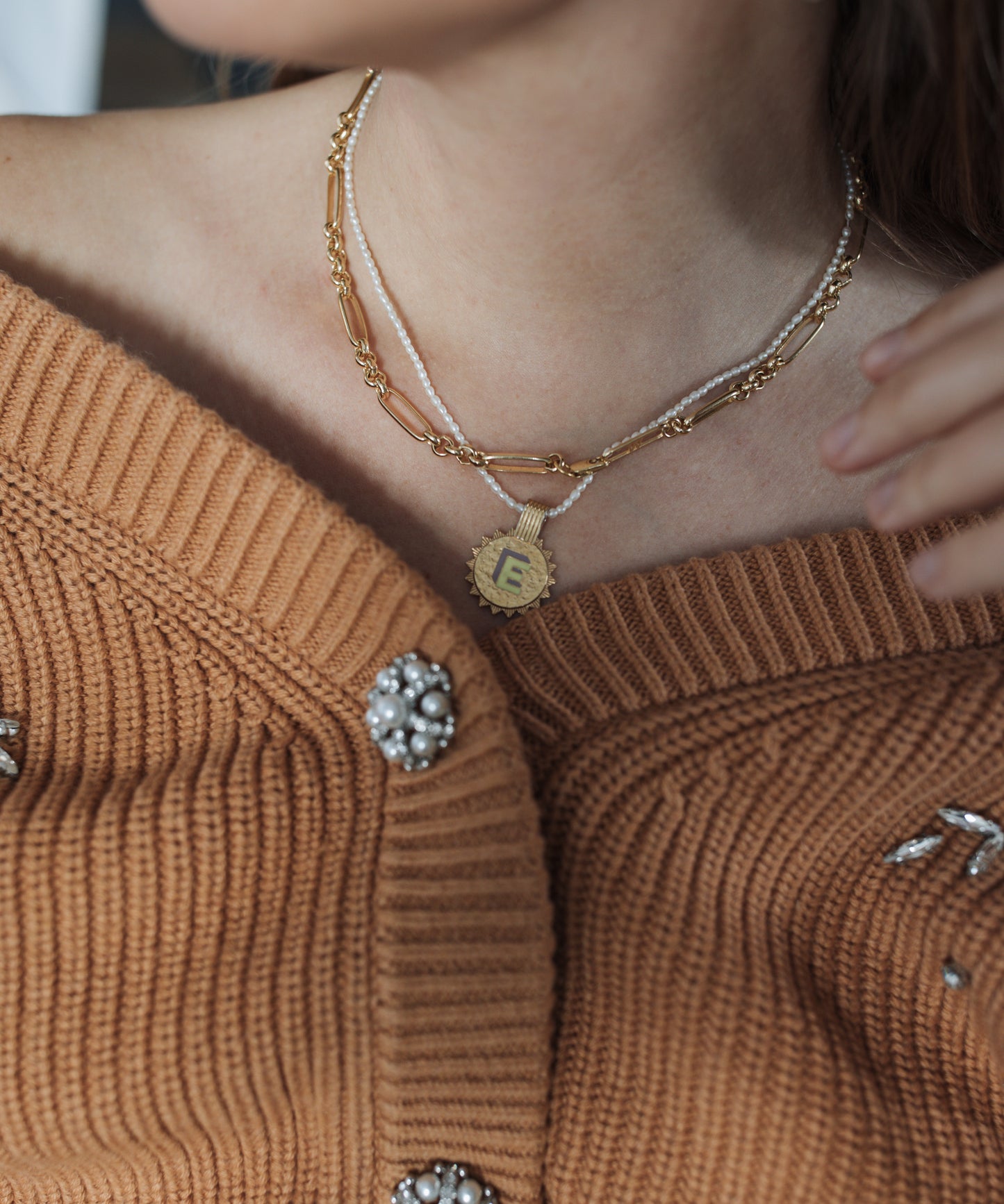 Close-up of a person wearing a brown knit cardigan with pearl embellishments and layered gold-plated necklaces, including WALD Berlin's Sample sale Say My Name Gold Necklace featuring an adjustable initial "E.