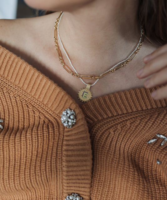 Close-up of a person wearing a brown knit cardigan with pearl embellishments and layered gold-plated necklaces, including WALD Berlin's Sample sale Say My Name Gold Necklace featuring an adjustable initial "E.