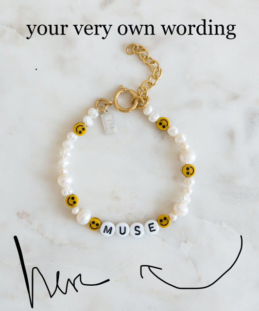 A WALD Berlin Personalized Bracelet featuring sweetwater pearls, cheerful yellow smiley faces, and letter beads spelling “MUSE” is displayed on a marble surface with customizable text above.