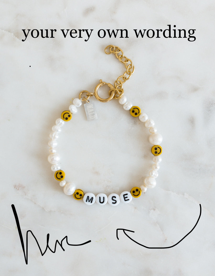 A WALD Berlin Personalized Bracelet featuring sweetwater pearls, cheerful yellow smiley faces, and letter beads spelling “MUSE” is displayed on a marble surface with customizable text above.